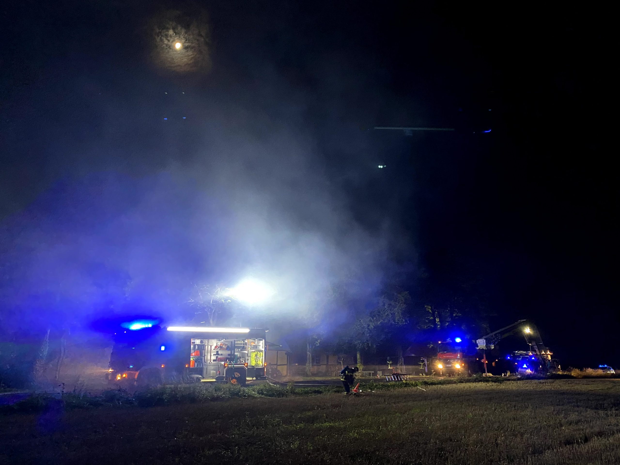 Brandbiler og redningskøretøjer oplyser et område med kraftige projektører under en natteaktion, omgivet af røg. Månen skinner svagt gennem en diset himmel, mens redningsarbejdere arbejder i baggrunden.