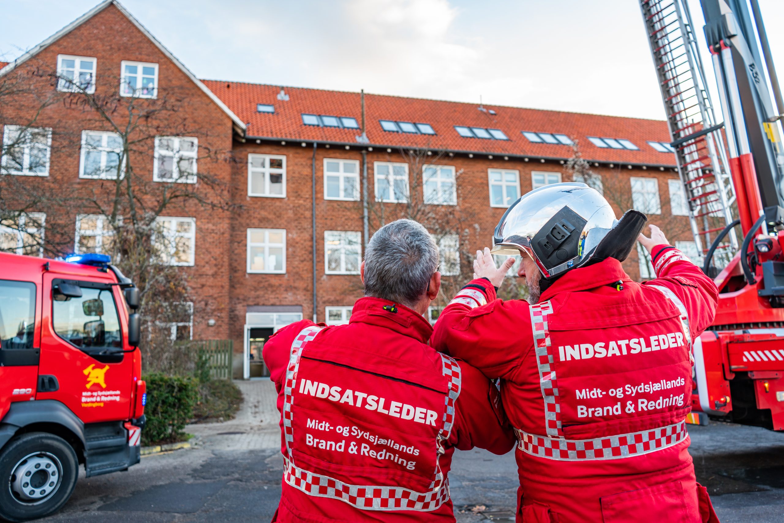 To indsatsledere fra Midt- og Sydsjællands Brand & Redning i røde uniformer vurderer en bygning under en redningsaktion. En brandbil og en stige ses i baggrunden ved siden af en murstensbygning